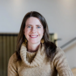 A white woman with  blue eyes and brown hair is wearing a tan cowl-necked sweater and smiling 